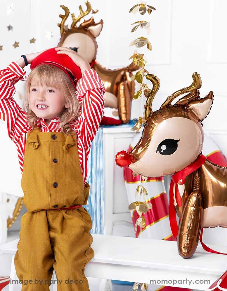A girl in candy cane red striped shirt sitting next to party deco's 24" reindeer foil balloon in a Christmas party