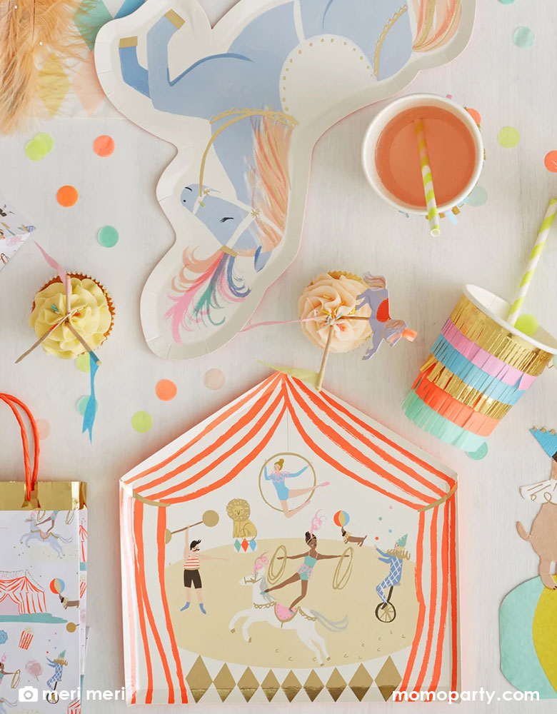 A party table filled with Momo Party's circus themed party supplies by Meri Meri including a big top tent shaped dinner plate, a circus themed party bag, a pony-shaped plate, a party cup with bright colored tassel decoration and some fun colorful confetti, which all makes a great inspiration for a kid's circus or carnival themed birthday party.