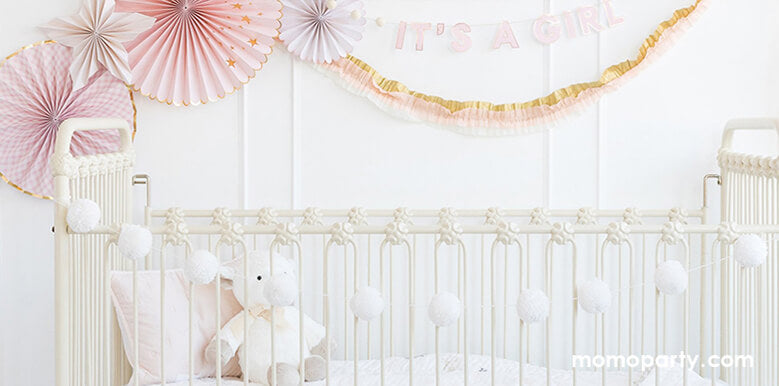 a pink white girl nursery room decoration with My Mind's Eye baby paper fans, it's a girl pink garland,  Cream Yarn Pom Pom Banner hanging on the crib, These super cute decoration so modern and cute for your newborn, nursery room decors.