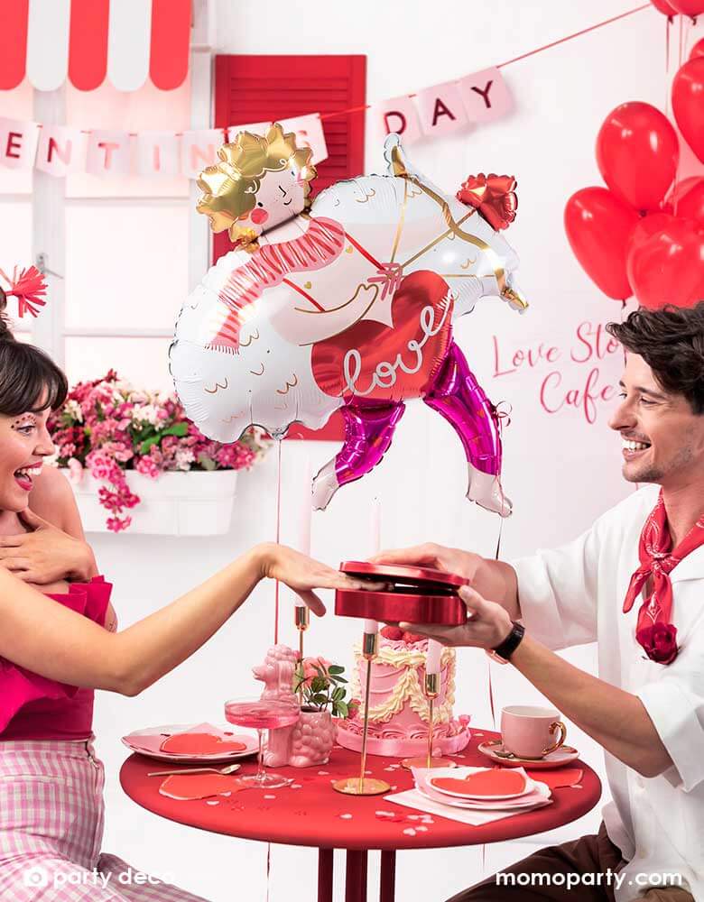 A loving couple at a dinner table enjoying a box of chocolate and pink cake to celebrate Valentine's Day. In the back the room is decorated with Momo Party's 32x34" Cupid shaped foil balloon and a bouquet of red heart shaped latex balloons, with a banner says Happy Valentine's Day in pink, it sets a romantic scene for a special Valentine's Day celebration.