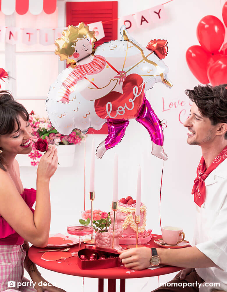 A loving couple at a dinner table enjoying a box of chocolate and pink cake to celebrate Valentine's Day. In the back the room is decorated with Momo Party's 32x34" Cupid shaped foil balloon and a bouquet of red heart shaped latex balloons, with a banner says Happy Valentine's Day in pink, it sets a romantic scene for a special Valentine's Day celebration.