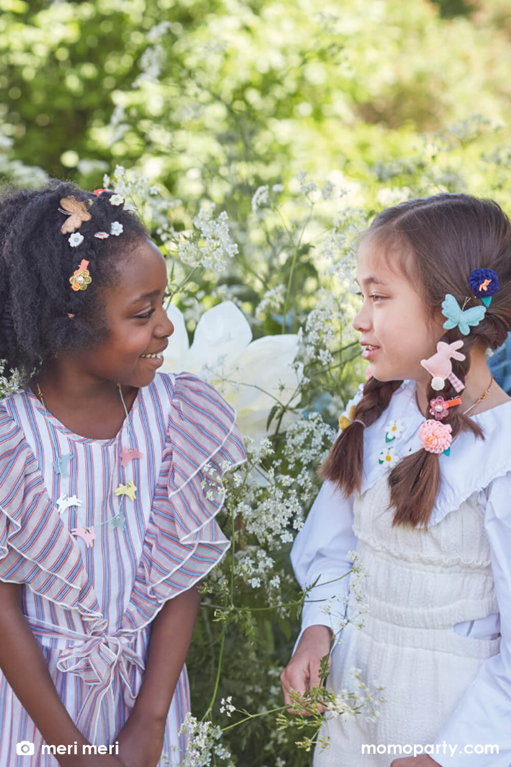 Two school aged girls chatting with each other in a beautiful garden with flowers blooming in the back. Both wore Meri Meri Easter themed accessories including bunny shaped felt hair clips, butterfly shaped felt hair chips and Meri Meri's acrylic leaping bunny necklace in 6 difference colors including  frosted blue, white, frosted pink, frosted mint, yellow and pink, with a gold tone chain and mint cord necklace. 