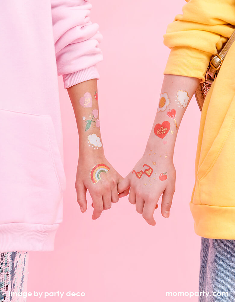 BFF Girls Interlocked fingers with lots of happy fun temporary tattoos like rainbow, sun, lollipop, heart cherry, daisy, heart apple on their arms.  