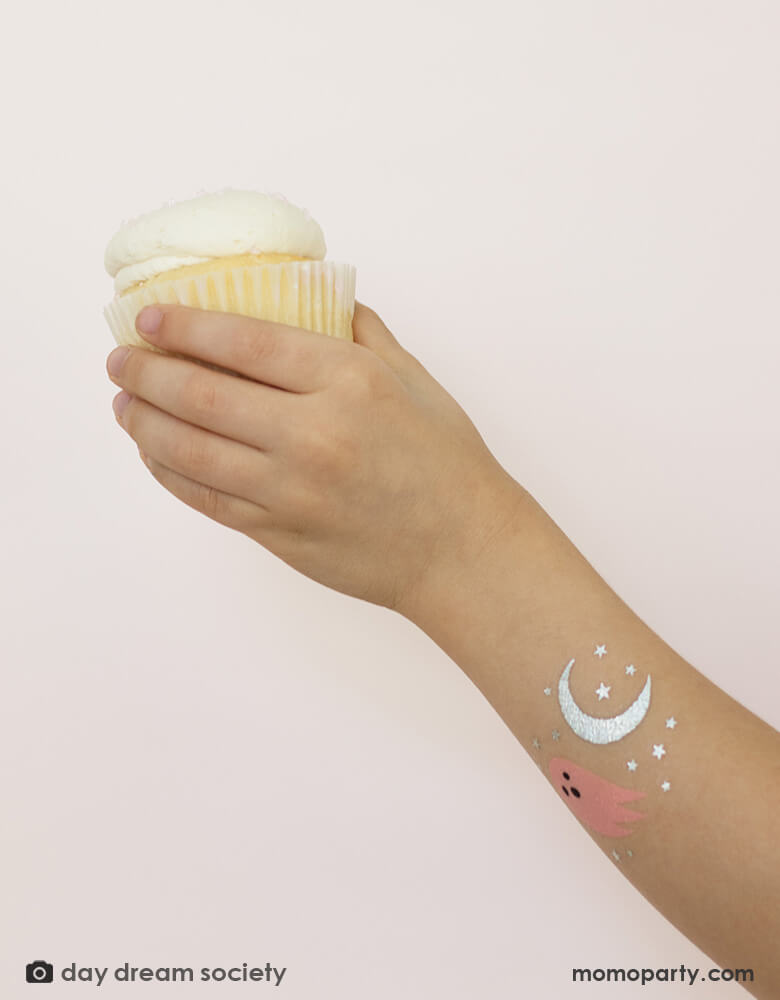 A girl holding a cupcake with her arm decorated with Daydream Society's Spooked temporary tattoos featuring ghost and moon designs 