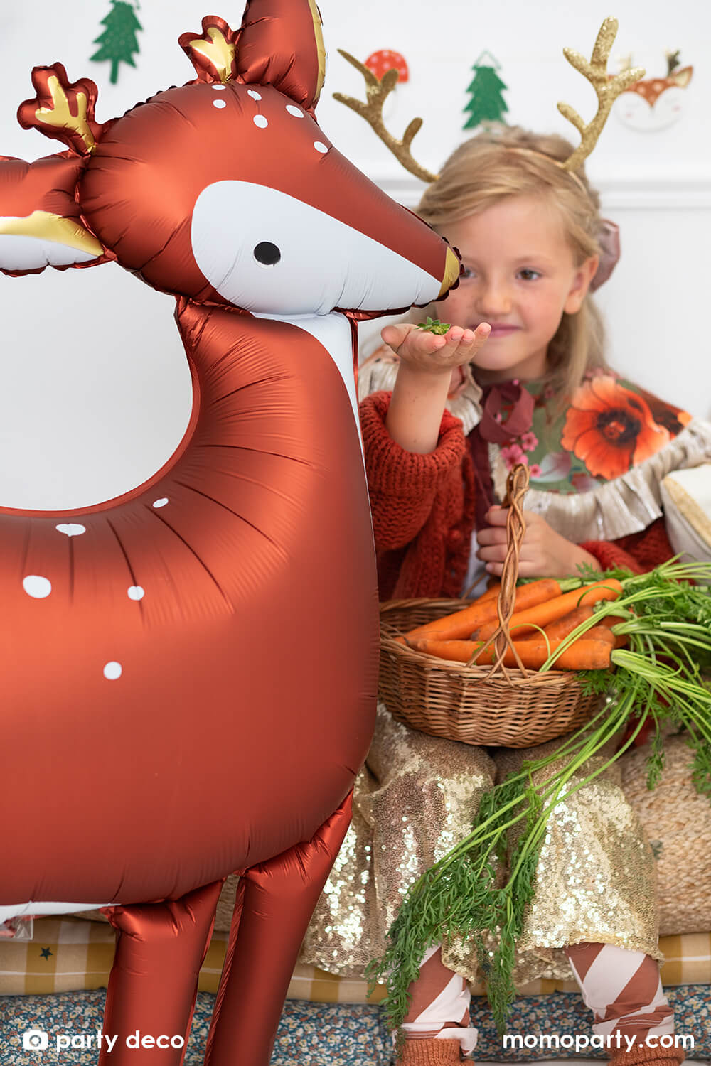 Christmas party fun with girl wearing a elope reindeer antlers headband, feeding a 40 inches large gorgeous deer shaped foil balloon in a modern room decorated with lots of holiday glitter garland, christmas tree and holiday themed foil balloons