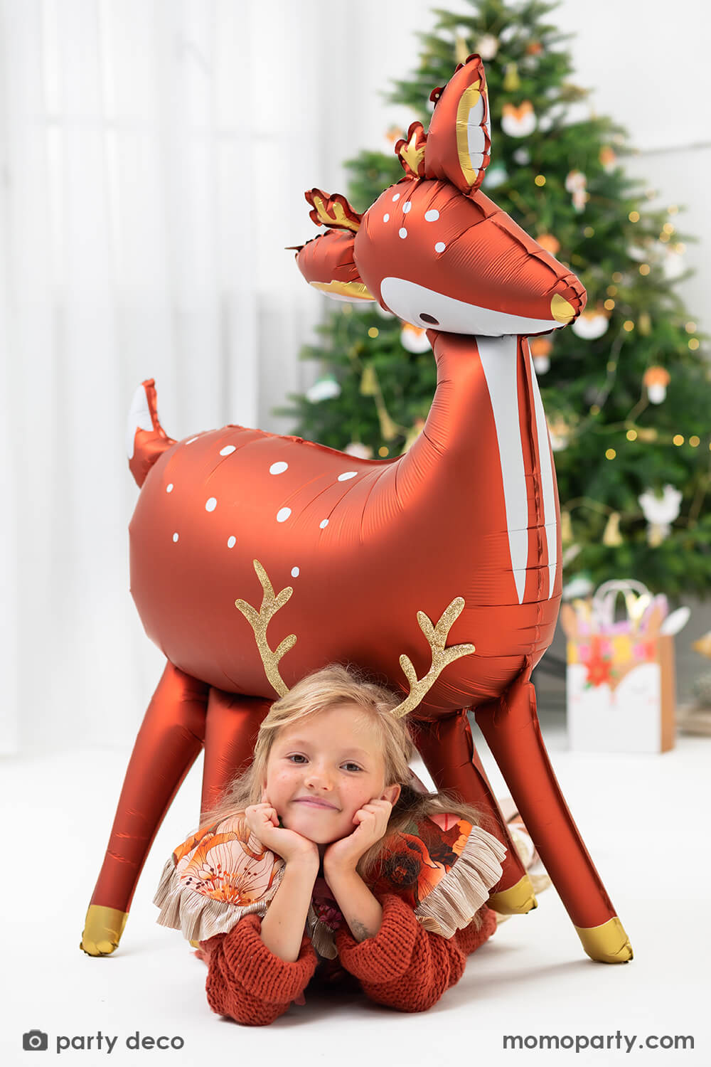 Holiday party fun with girl wearing a elope reindeer antlers headband, playing under a 40 inches large gorgeous standing deer shaped foil balloon in a modern clean room decorated with lots of holiday garland, christmas tree and holiday themed foil balloons