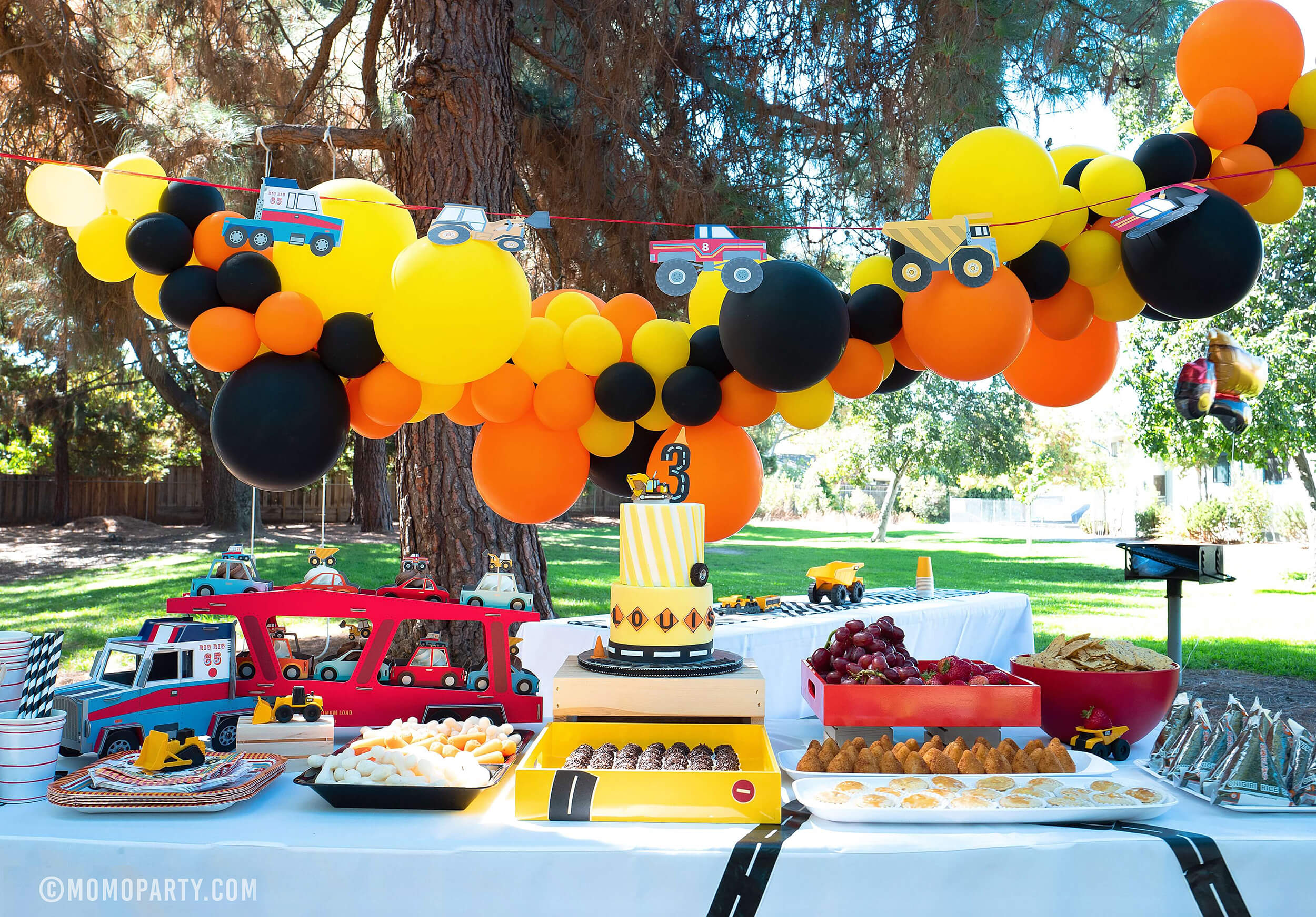 Dig-In Construction Outdoor Party table set up with balloon garland, Truck Garland, Truck Cupcake stand, Cakes and food