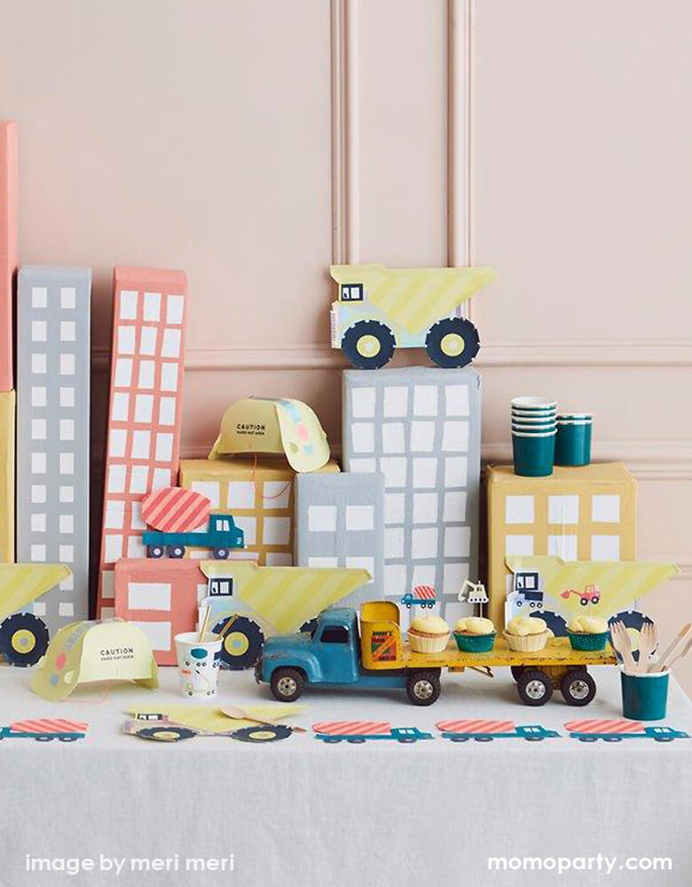 Kid Dig In Construction party table with lot of cupcakes with construction cupcake kit topper on a vintage truck toy, with Meri Meri Dumper Truck Plates, Construction Party cups and Construction truck napkins around, and paper made pastel color building as backdrop. what a Fun and modern party set up idea for a kid's construction themed birthday party, Dig in birthday party, big construction vehicle lover, boy's birthday party, kid's birthday party