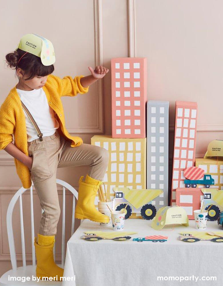 A girl wearing a yellow Construction Party Hat, a yellow sweater, she standing on a chair lifting her yellow rainboot in a Kid Dig In Construction party table. There are Meri Meri Dumper Truck Plates, Construction Party cups and Construction truck napkins, and paper made pastel color building as backdrop. what a Fun modern partware for a kid's construction themed birthday party, Dig in birthday party, big construction vehicle lover, boy's birthday party, kid's birthday party
