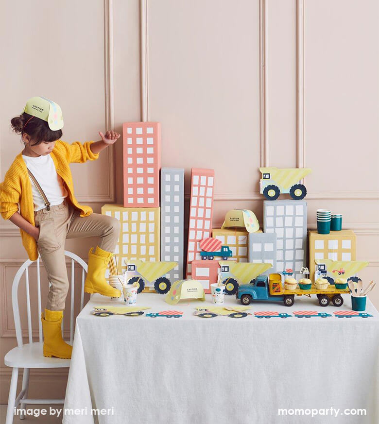 A girl wearing a Construction Party Hat, yellow sweater and yellow rain boots, standing on a chair next to a party table. There are Meri Meri Dumper Truck Plates, Construction Party cups and Construction truck napkins, cupcakes with vehicle toppers from Construction Cupcake Kit and colorful cardboard made buildings as backdrop. what a Fun modern partware for a kid's construction themed birthday party, Dig in birthday party, big construction vehicle lover, boy's birthday party, kid's birthday party