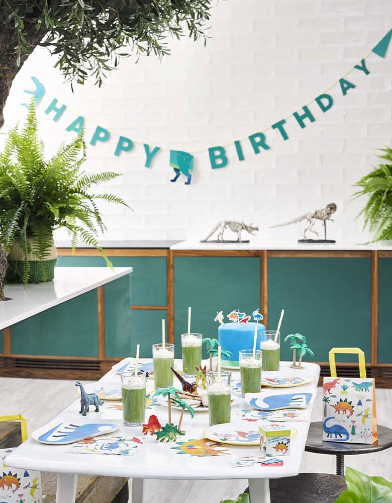 A kid's dinosaur themed birthday party featuring Talking Tables' Dinosaur Collection including Happy Birthday Garland in green with dinosaur's head and tail on the sides of the banner hung on the wall and dinosaur themed table tops on the party table in a jungle-like set up