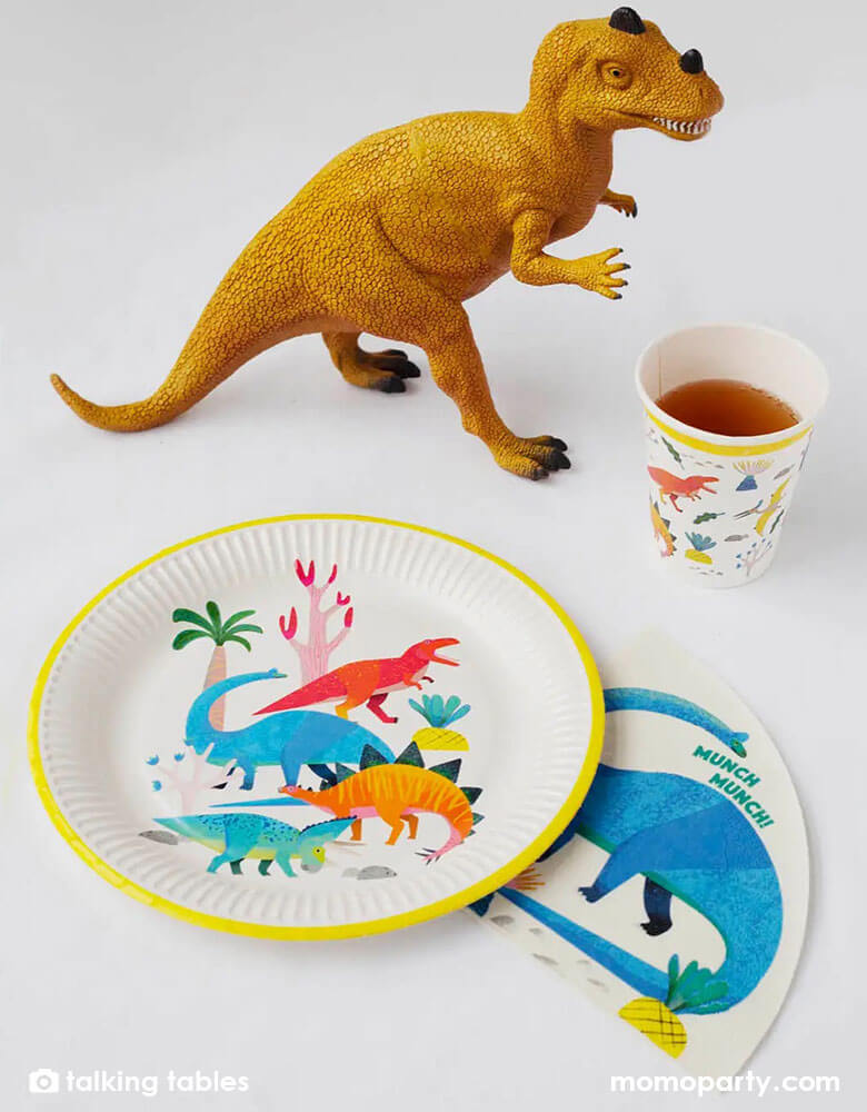 A table filled with talking tables 9 inch dinosaur round plates dinosaur party cups and dinosaur napkins with a toy dinosaur figure 