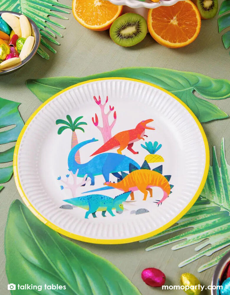 A kid's jungle dinosaur themed party table with talking table's 9 inch dinosaur round plates with palm tree leaves decorations around it and tropical fruit with candies in bright colors