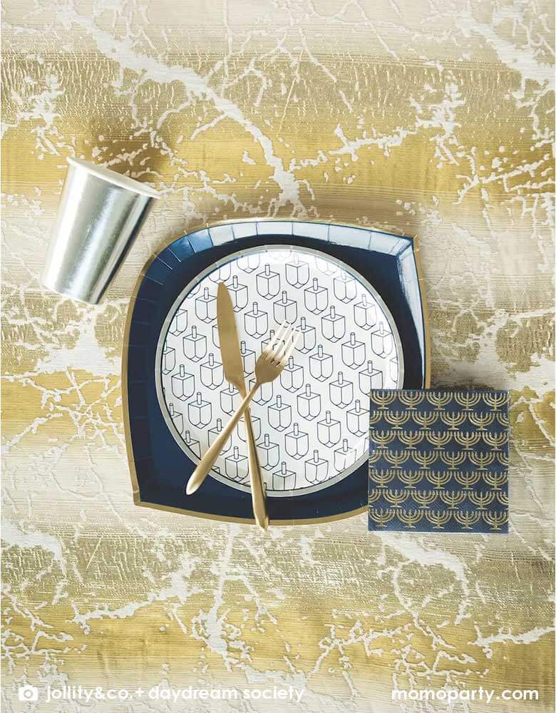 A modern and elegant Hanukkah tablescape featuring a silver party cup, a large navy napkins with gold menorah and Momo Party's 9" round plate by Jollity & Co. featuring silver foil rim and navy printed Dreidels.