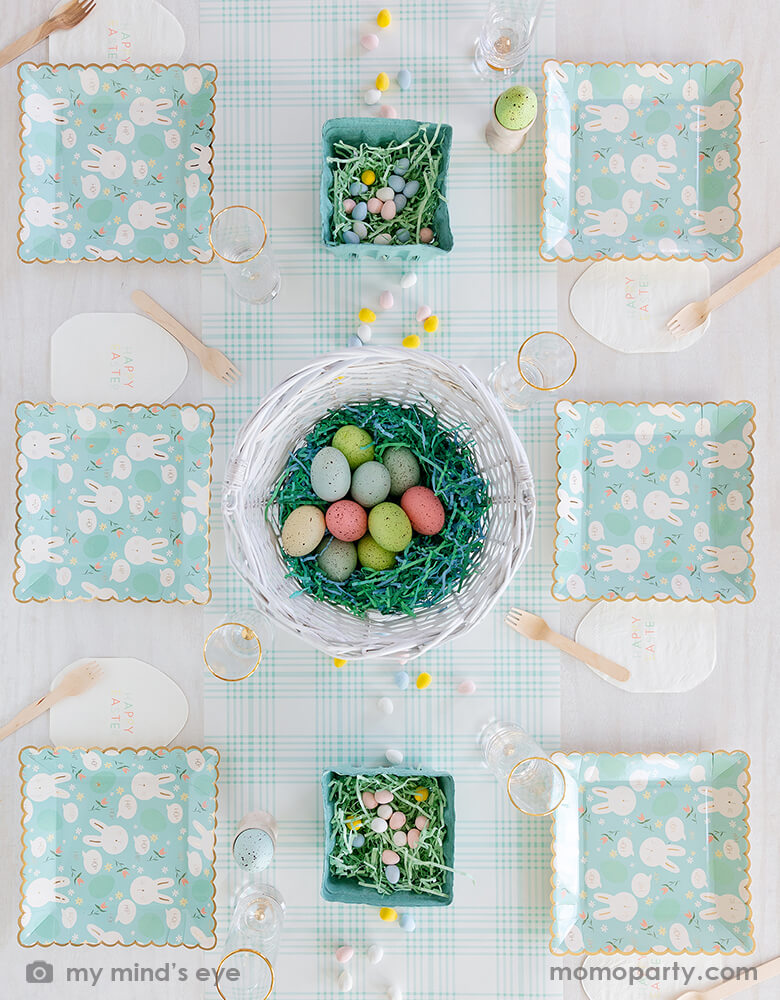 Top view of Easter party tabletop for 6 people covered with 6 sets of My Mind's Eye - Scattered Bunny Scallop Paper Plates with Easter Egg Shaped Napkins, Wooden Forks and Drinks, With a white basket full of colorful eggs in the middle of the table and 2 small green baskets filled with mint shredded eggs on a Blue Plaid Paper Table Runner as a centerpiece, these fresh party supplies with a modern and fun design are perfect for Easter celebrations, easter party and spring party