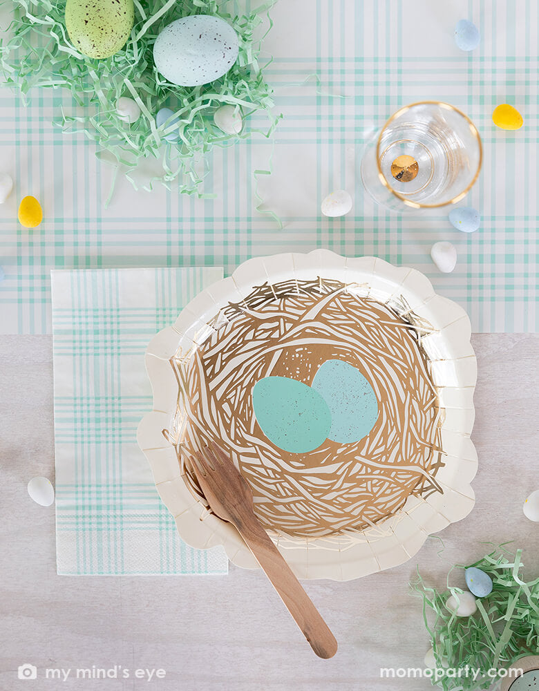Top view of Easter party table top covered with My mind's eye -Nest shaped paper plates with Blue Plaid Guest Towel Paper Napkins and wooden fork, all on a blue checkered paper table runner, surrounded by colored eggs on mint shredded paper filling like a bird's nest and a glass of drink. These fresh partyware with modern fun designs are perfect for Easter celebrations
