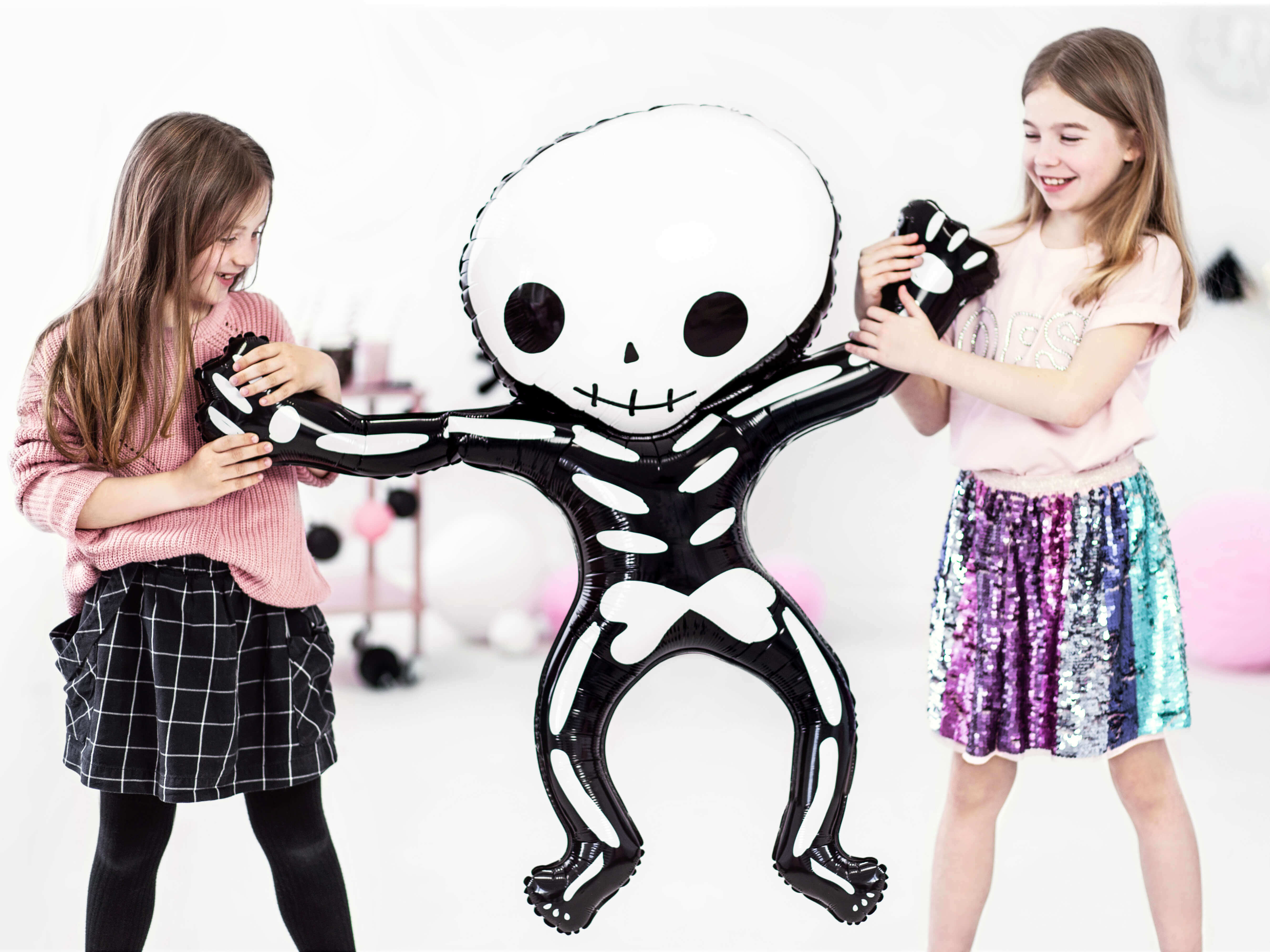 2 Girls holding a Party Deco - Halloween funny Skeleton shape Foil Mylar Balloon in a pink halloween party. This fun skeleton foil balloon like a kid size, also perfect for Kid-Friendly modern spooky halloween party, trick-or-treating halloween party, nightmare before christmas party, witch themed party and all halloween related celebrations