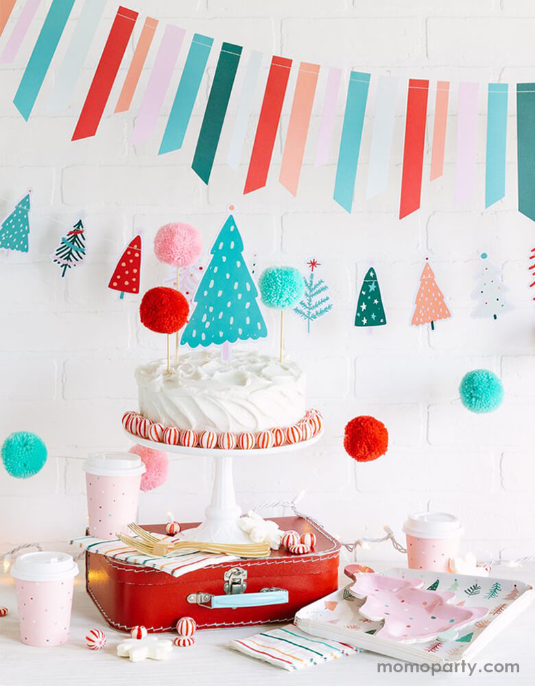 My Minds Eye & Oui Party Collab Christians party Dessert table set up with a buttercream white cake with pom pom cake topper and peppermint around, with a cake stand on top of a red vintage suitcase. a set of FA LA LA 9" FROSTING TREE PLATEs with FA LA LA CHRISTMAS 9" TREES PLATEs on the side, a  6ft Fa La La Christmas Tree Banner in cheery pastel colors and FA LA LA TICKER TAPE BANNER hanging on the wall as decoration for a pastel cute cozy christmas party