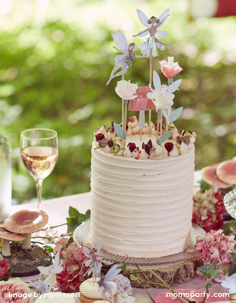 A dreamy outdoor fairy garden party look of a beautiful cream butter cake with Meri Meri Fairy Cake Topper. there are 3 fairies, a toadstool and flowers topper on top of it, a glass of rose, lots of flowers around, cupcake with fairy cupcake topper