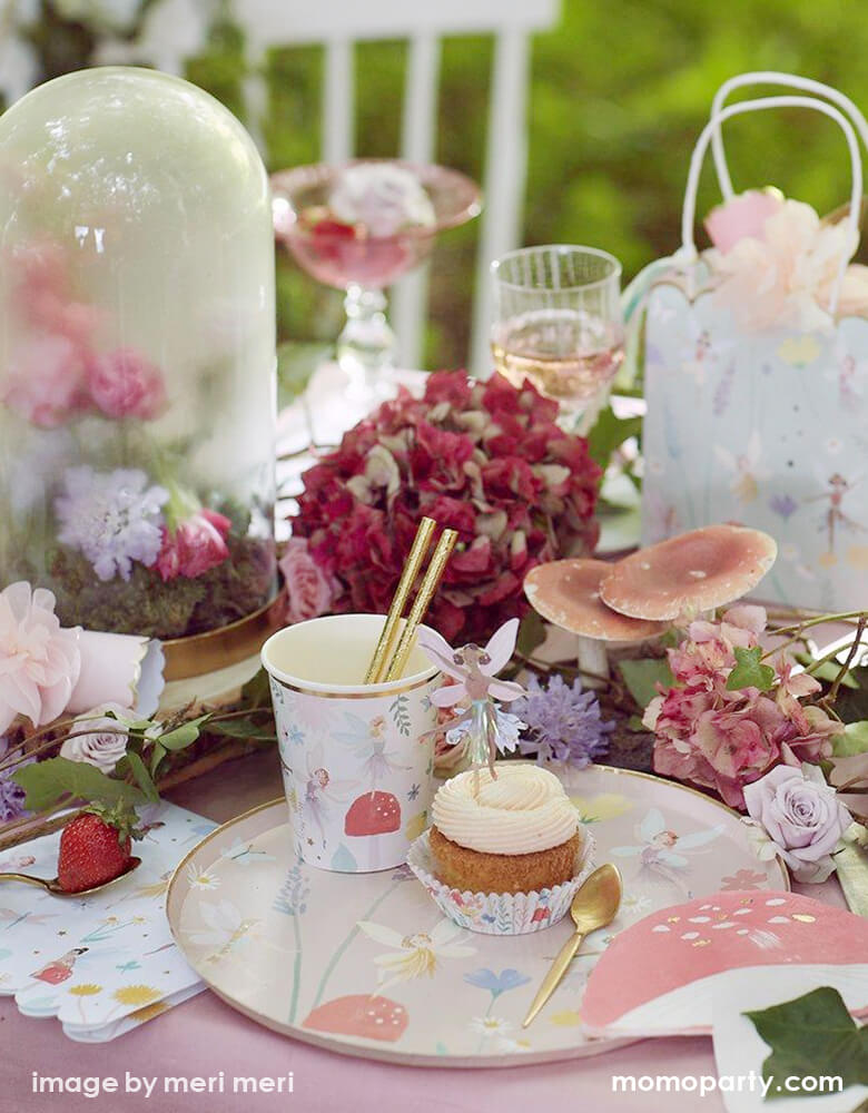 A outdoor Fairy themed birthday party table close up look of a cupcake with Fairy cupcake topper, and a fairy paper party cups with a gold foil straw on top of a Meri MerFairy Dinner Plate, lots of flowers, mushrooms, leaves, strawberries around as decoration, such a dreamy modern look fairy party for kids, Tea party party with mom and daughter, perfect idea for a girl's birthday party, garden party, spring party, summer party