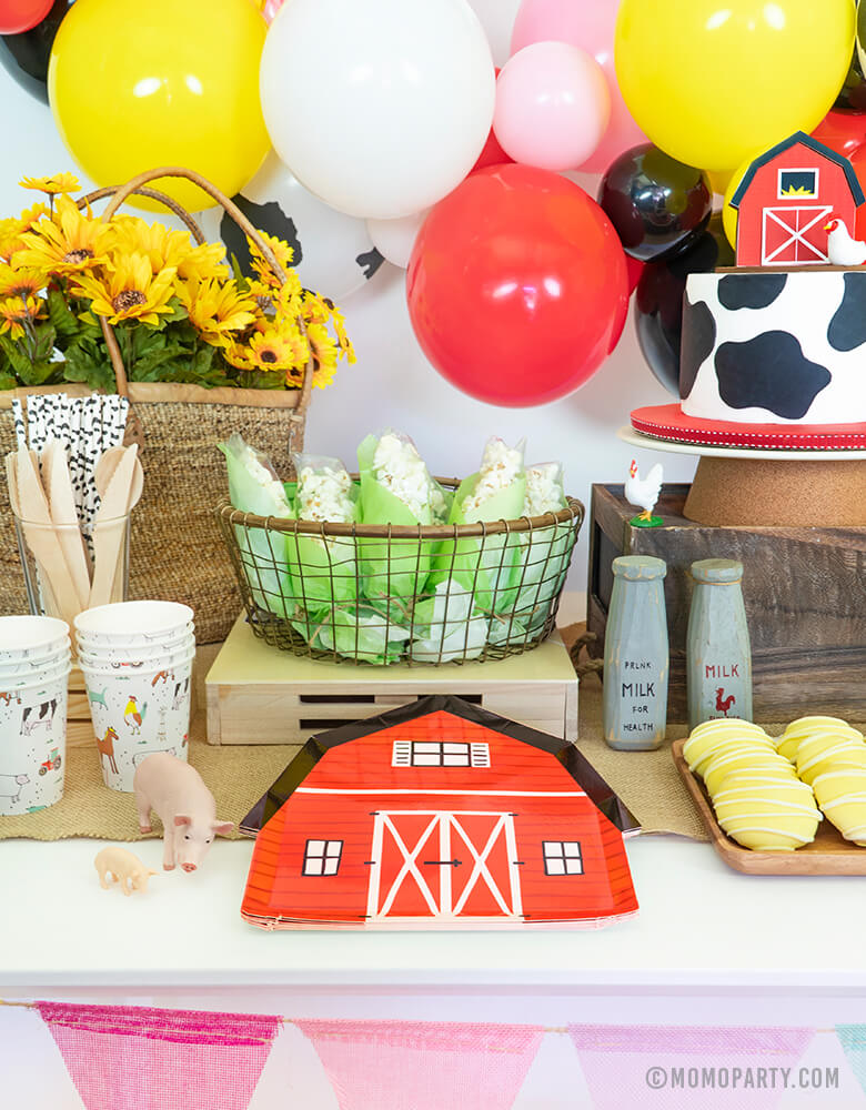 Dessert Table for Barn yard, Farm Animals Birthday Party with Barn house party plate, popcorn corn basket, cow cake, farm animals paper cups, cookies, flowers in the basket, and balloon garland on the back