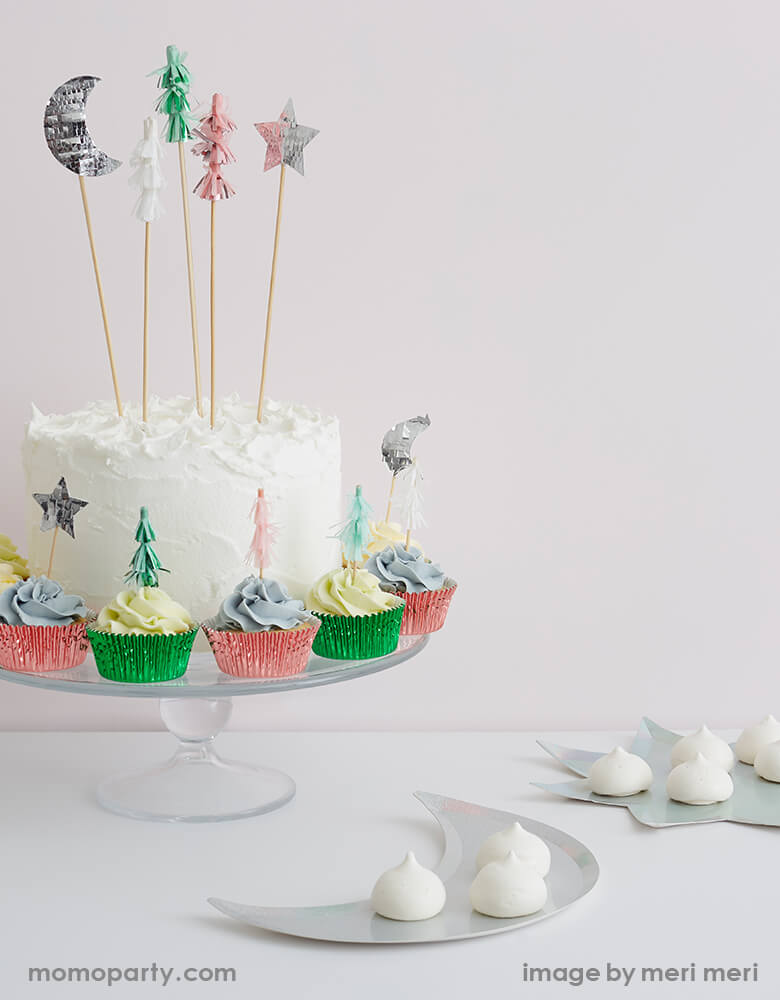 A cake with whipped cream and Festive Tree cake toppers, surrounded by a Set of cupcakes topped with Meri Meri Festive-Tree-Cupcake-Kit featuring Christmas tree, moon and star toppers in pink silver and mint foil