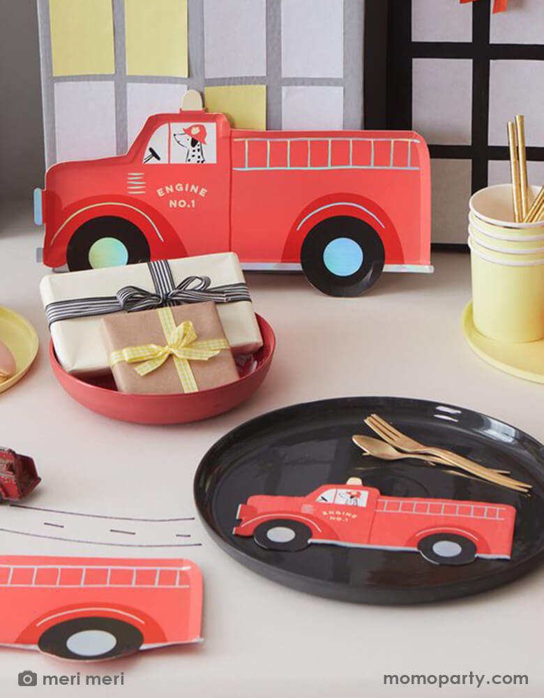 An adorable fire truck fire fighter themed party table with Meri Meri's fire truck themed party plates and napkins, along with pale yellow and red solid color plates and tumblers. On the table there was a vintage firetruck toy riding on the table drawn with roads and some gift boxes wrapped with simple bows in modern colors of red and pale yellow, in the back there were cardboard made city buildings mimic the city scene. This is a perfect inspiration for kid's fire truck / firefighter themed birthday party
