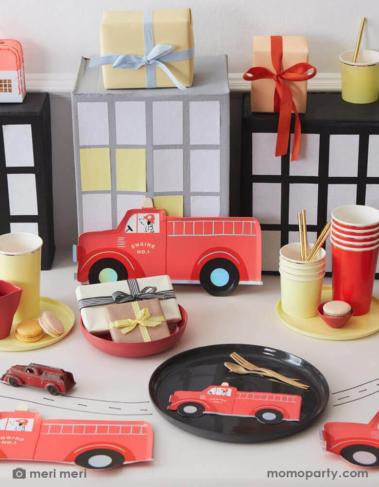 An adorable fire truck fire fighter themed party table with Meri Meri's fire truck themed party plates and napkins, along with pale yellow and red solid color plates and tumblers. On the table there was a vintage firetruck toy riding on the table drawn with roads and some gift boxes wrapped with simple bows in modern colors of red and pale yellow, in the back there were cardboard made city buildings mimic the city scene. This is a perfect inspiration for kid's fire truck / firefighter themed birthday party