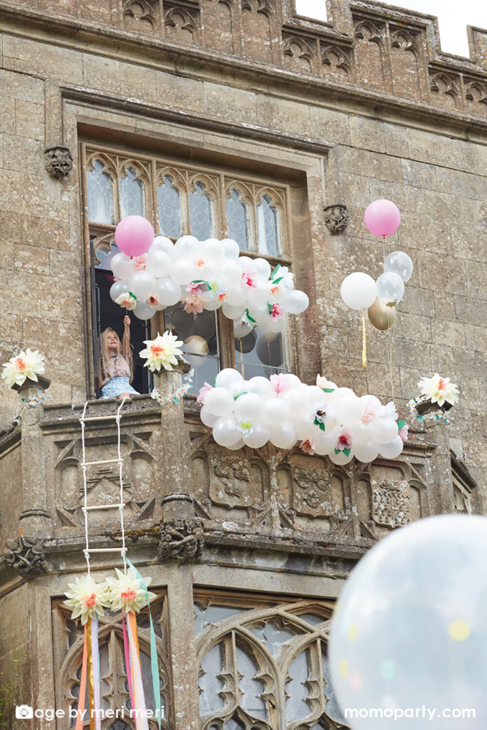 A girl standing on the balcony of a whimsical castle looking building that is decorated with Meri Meri Floral Balloon Garland made with pearl balloons and paper flowers which are crafted from pink, peach, yellow and white tissue paper with golden foil centers.