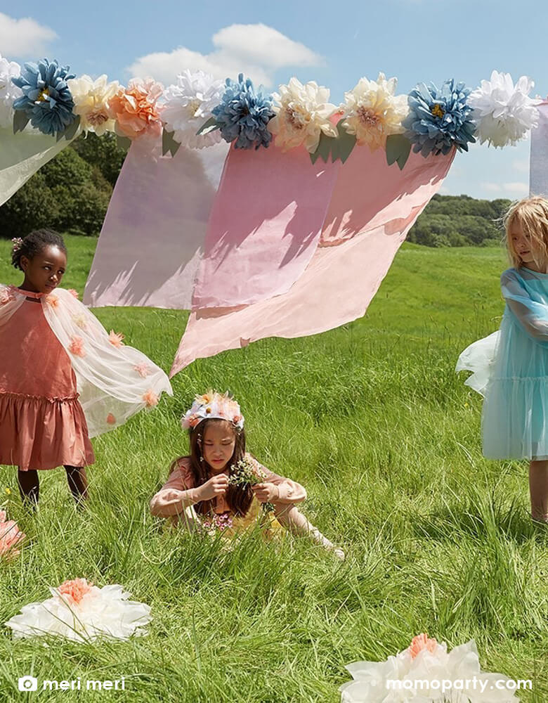 An outdoor spring themed party in an open lawn for girls decorated with Meri Meri's 8.5 ft pre-strung giant flowers in bloom garland featuring beautiful flowers in peach, white, dusky blue,cream, yellow and dusky pink, made with tissue paper, along with green leaves.