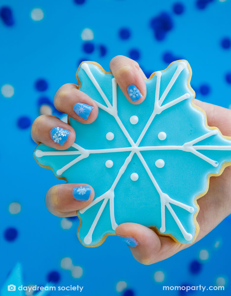 kid holding a snowflake shaped cookie with her light blue colored fingernail color decorated with Frosted Nail Stickers from Jollity & Co Party Boutique - Daydream society- Frosted collection. Featuring variety colors of snowflakes in morden illustrations with some iridescent details, These frosted nail stickers are perfect for stocking stuffers and goodie bags, activity for a frozen themed birthday party, winter wonderland party, holiday fun activity for mother daughter time.