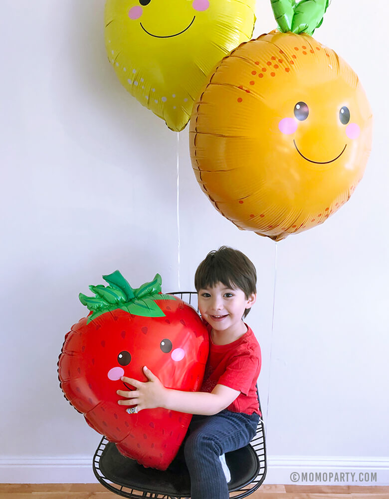 boy sitting under lemon and orange foil balloons and hugging Strawberry Foil Mylar Balloon
