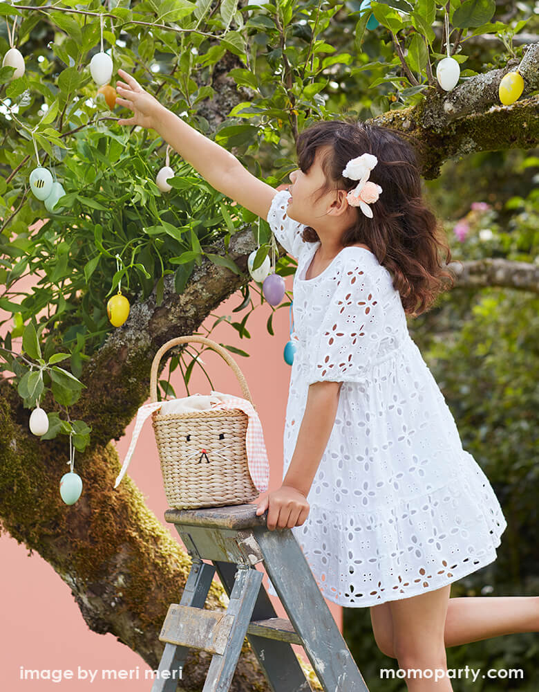 A girl wearing a cute white dress, standing on the ladder with Meri Meri Gingham Bunny Straw Easter Basket, picking easter eggs from tree,  Meri Meri - Gingham Bunny Straw Easter Basket.