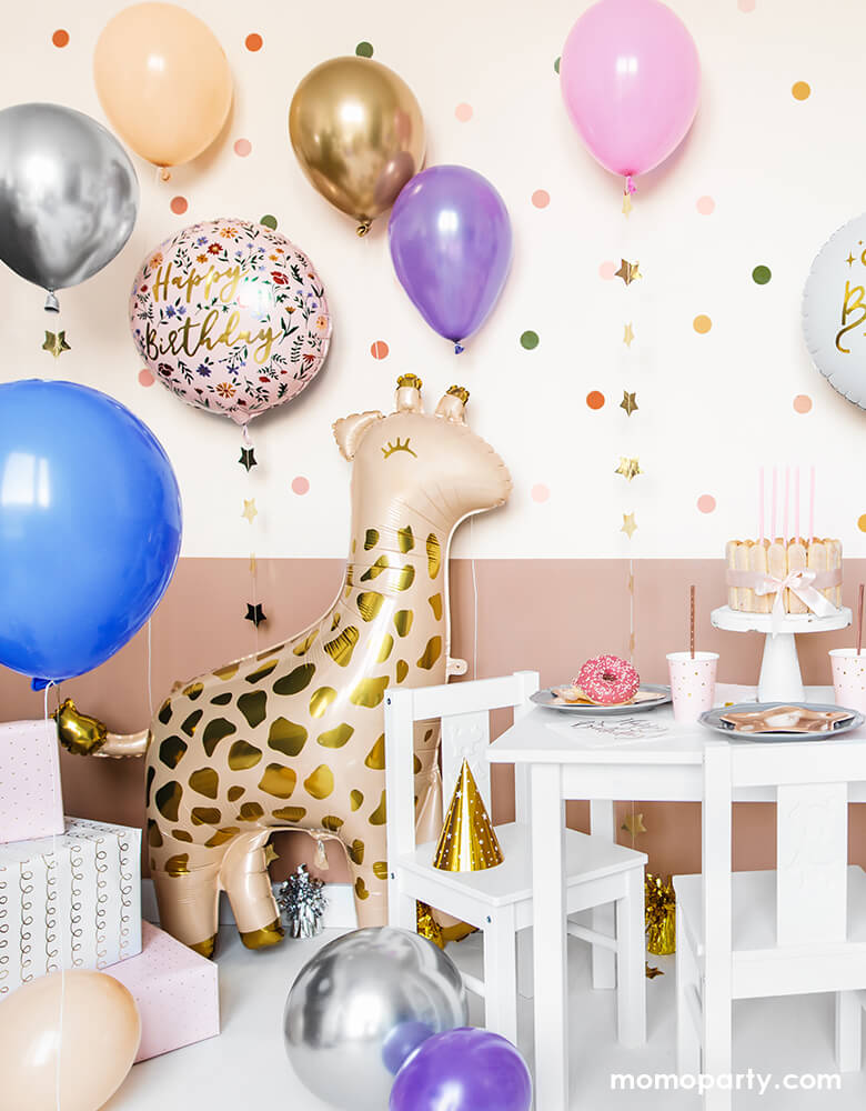 A loving birthday celebration at home, fill with lots of gold, pink, blue, silver, blush latex balloons, Junior Floral Happy Birthday Foil Balloon, Gifts on the conner of the room next to a 40 inch Giant Smelling Giraffe Foil Mylar Balloon by Party Deco standing next to the kids desk and chairs. there are cake, donut, sweets, and party ware on the kid's table. Ready for the little one's safari or zoo themed birthday celebration