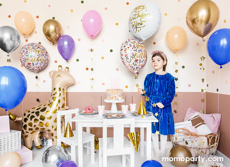 A loving birthday celebration at home, fill with lots of gold, pink, blue, silver, blush latex balloons, a girl wearing a blue dress holding a Junior Floral Happy Birthday Foil Balloon, Gifts on the conner of the room next to a 40 inch Giant Smelling Giraffe Foil Mylar Balloon by Party Deco standing next to the kids desk and chairs. there are cake, donut, sweets, and gold party hats, and party ware on the kid's table. Ready for the little one's safari or zoo themed birthday celebration
