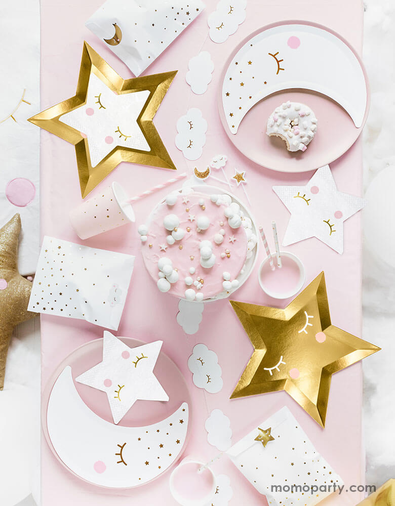 A flatlay shot of a baby pink table setup for a sweet baby shower filled with sweet Star themed party goods including die cut gold star plates, little star napkins in white, moon shaped plates in white with pink party cups and party favor bags
