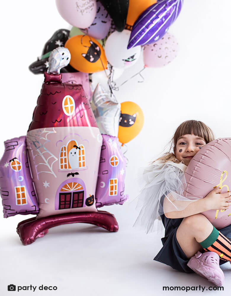 Girl in front of Party Deco 35x46 inches haunted house shaped standing foil balloon with cute ghost illustrations in pink and iridescent details, a perfect decoration for a girly pink Halloween party