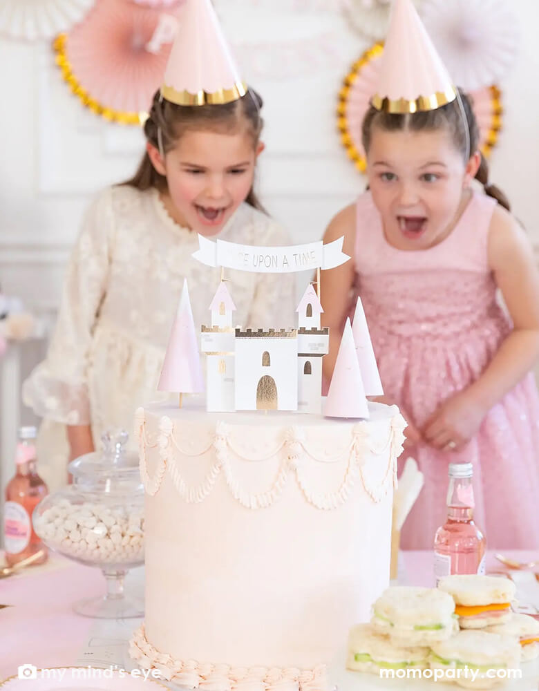 A group of girls dressed up as princesses in princess party hats and star wands standing in the back of a gorgeous princess themed pink birthday cake decorated with My Mind's eye cake topper set featuring pieces of castles, mountains, and "once upon a time" banner