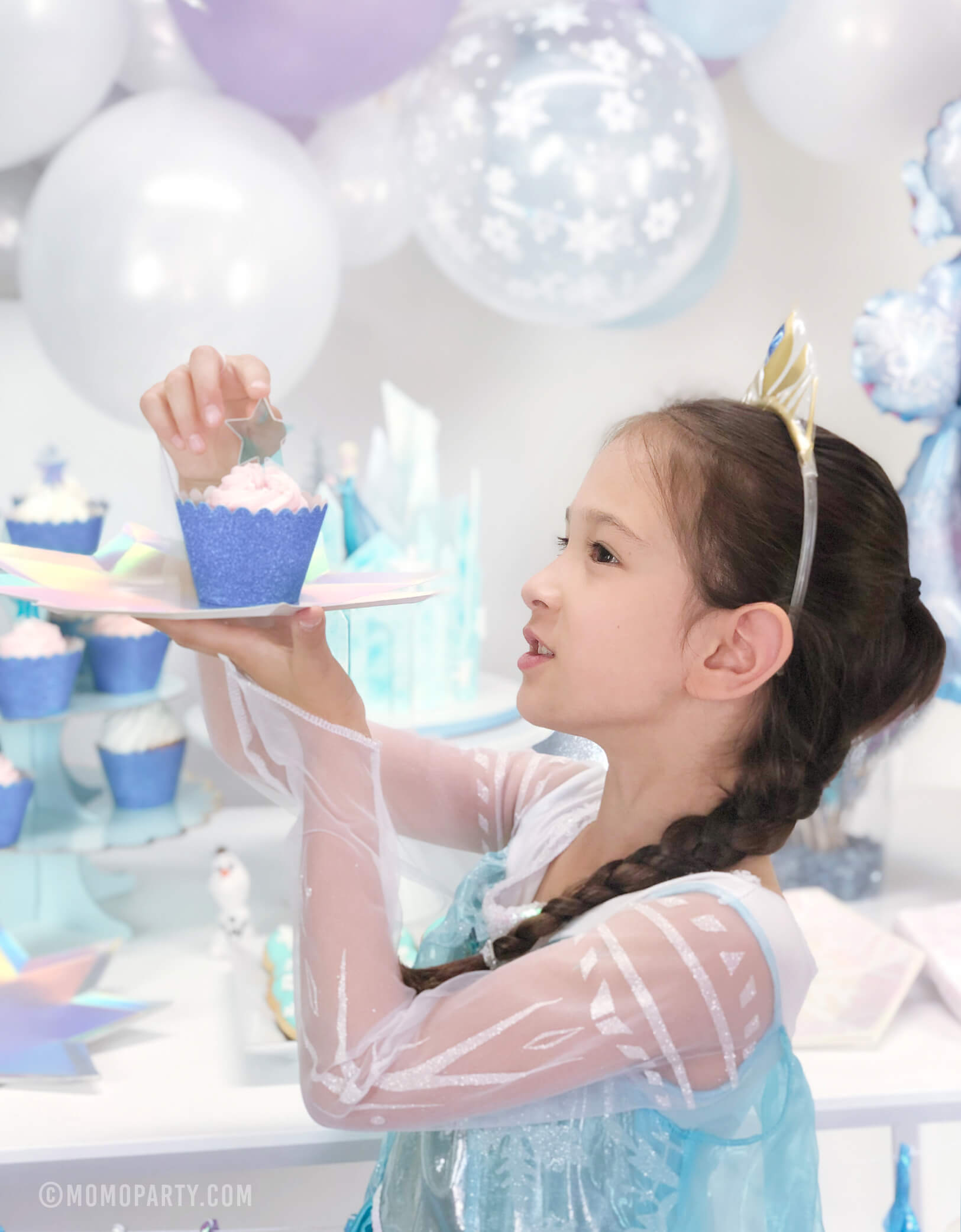 A girl wearing Elsa Princess costume holding a cupcake with Glitter Ice Blue Cupcake Wrapper on a star plate in her Frozen themed birthday party 