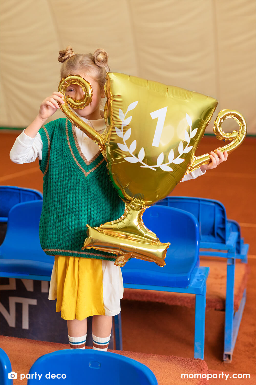A little girl in school uniform holding party deco's Party Deco's 28" gold cup trophy shaped foil balloon in glossy material with number 1 on it, celebrating her win in a tennis court