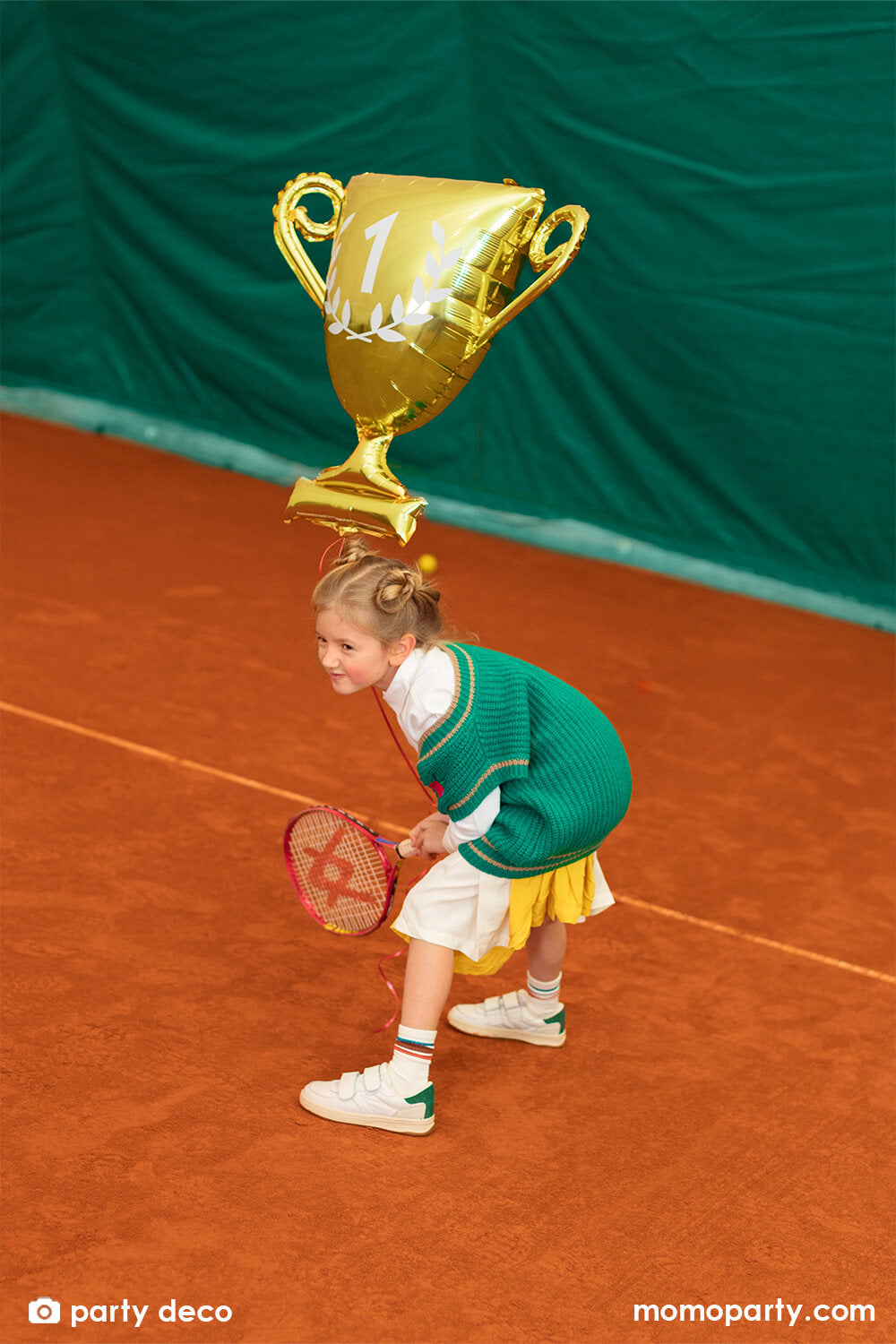 A little girl in school uniform holding her tennis racket in a gam with Party Deco's Party Deco's 28" gold cup trophy shaped foil balloon in glossy material with number 1 on it, celebrating her win in a tennis court