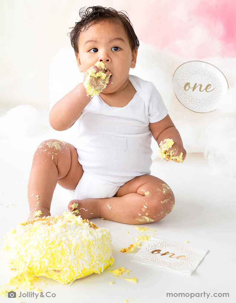 A baby's boy's first birthday smash cake celebration with Jollity 7-inch onederland gold round dessert plate with gold script "one" on it and gold foil confetti illustration around it with coordinated Jollity 5 inch onederland small napkins spread around