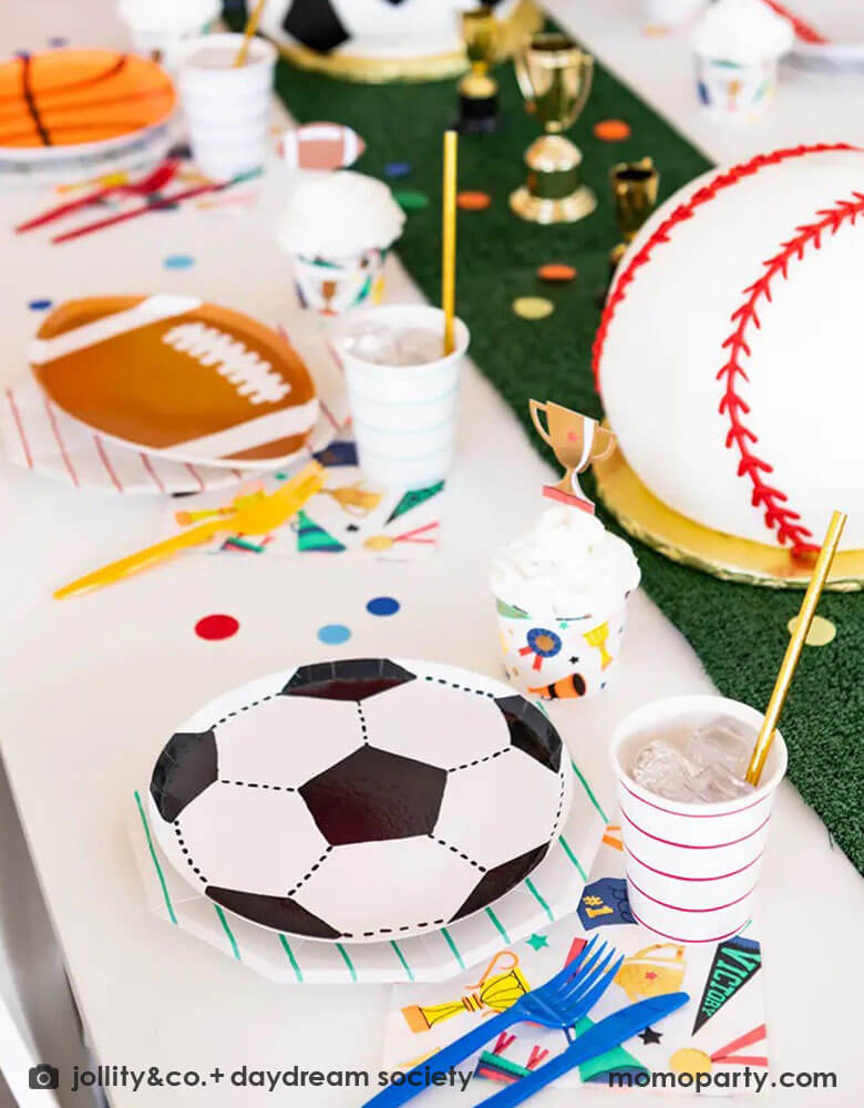 A fun party table filled with Momo Party's sports themed party tableware by Daydream Society including die-cut soccer ball shaped plates, football shaped plates, basketball shaped plates with mint and red striped plates and cups, plus some good sport large napkins featuring bright colors and fun illustrations of sports elements. In the center of the table there's a faux grass table runner and baseball shaped birthday cake, all makes great inspirations for kid's sport themed parties.