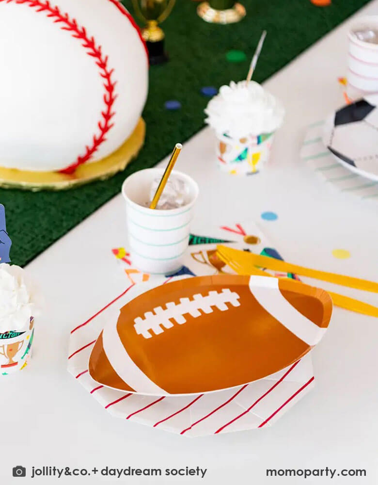 A fun party table filled with Momo Party's sports themed party tableware by Daydream Society including die-cut soccer ball shaped plates, football shaped plates, basketball shaped plates with mint and red striped plates and cups, plus some good sport large napkins featuring bright colors and fun illustrations of sports elements. In the center of the table there's a faux grass table runner and baseball shaped birthday cake, all makes great inspirations for kid's sport themed parties.