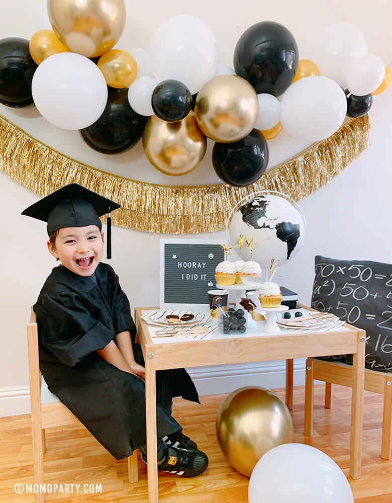 happy little kindergarten graduator wearing a black graduation gown celebrate at home with a modern graduation party look, which has black, gold and silver balloon garland, Gold Tinsel Fringe Garland on the wall, black stripe plates, napkins, Assorted Gold Dipped Cups, b&w globe, letter board with "hooray i did it" sign, cupcakes, black gum balls on a kid desk