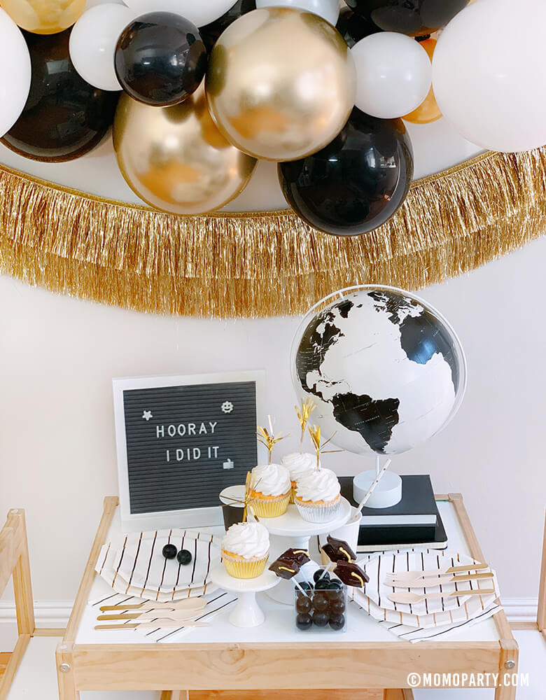 front look of a modern graduation party at home with black, gold and silver balloon garland, Black Graduation Cap Foil Balloon, Gold Tinsel Fringe Garland on the wall, black stripe plates, napkins, Assorted Gold Dipped Cups, b&w globe, letter board with "hooray i did it" sign, cupcakes, black gum balls on a kid des
