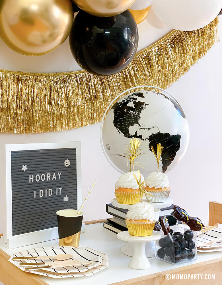 Modern graduation party at home idea with black, gold and silver balloon garland, Black Graduation Cap Foil Balloon, Gold Tinsel Fringe Garland on the wall, black stripe plates, napkins, Assorted Gold Dipped Cups, b&w globe, letter board with "hooray i did it" sign, cupcakes, black gum balls on a kid desk