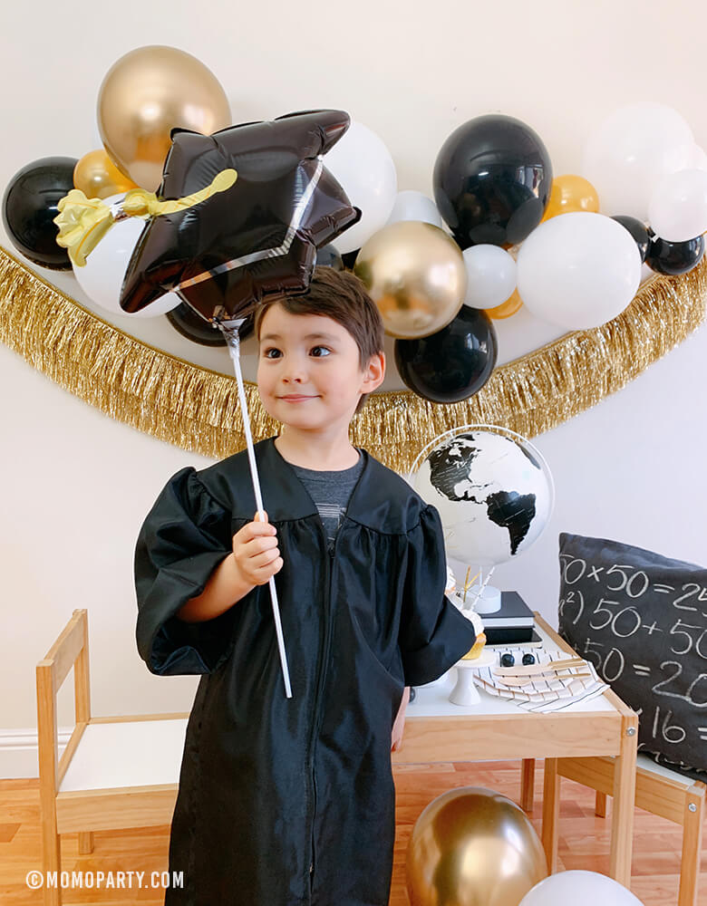 a kindergarten boy wearing a black graduation gown, holding a Black Grad Cap Mini Foil Balloon, celebrate at home with a modern graduation party look, which has black, gold and silver balloon garland, Gold Tinsel Fringe Garland on the wall, black stripe plates, napkins, Assorted Gold Dipped Cups, b&w globe, letter board with "hooray i did it" sign, cupcakes, black gum balls on a kid desk