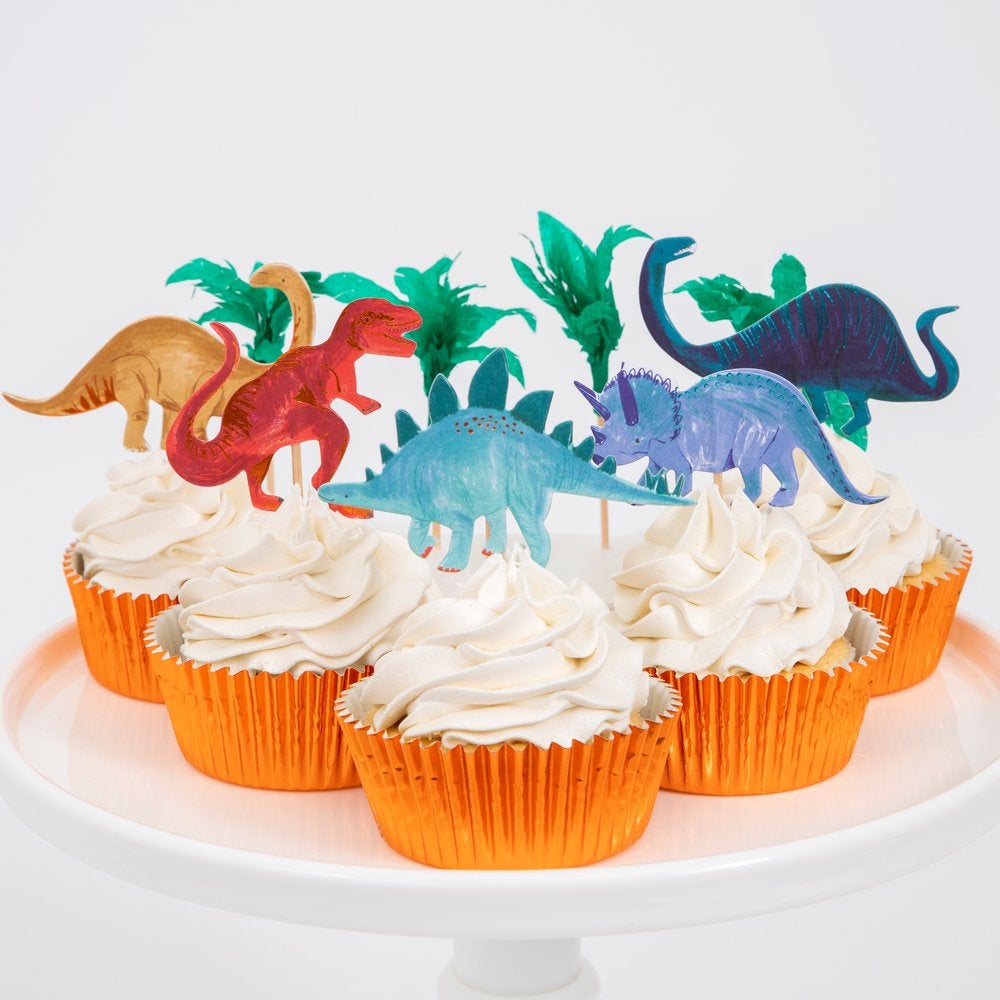 Cupcakes in a white cake stand, decorated with Meri Meri Dinosaur Kingdom Cupcake Kit, toppers featuring in the design of t-rex, Triceratops and paper palm trees and shimmering copper foil cases