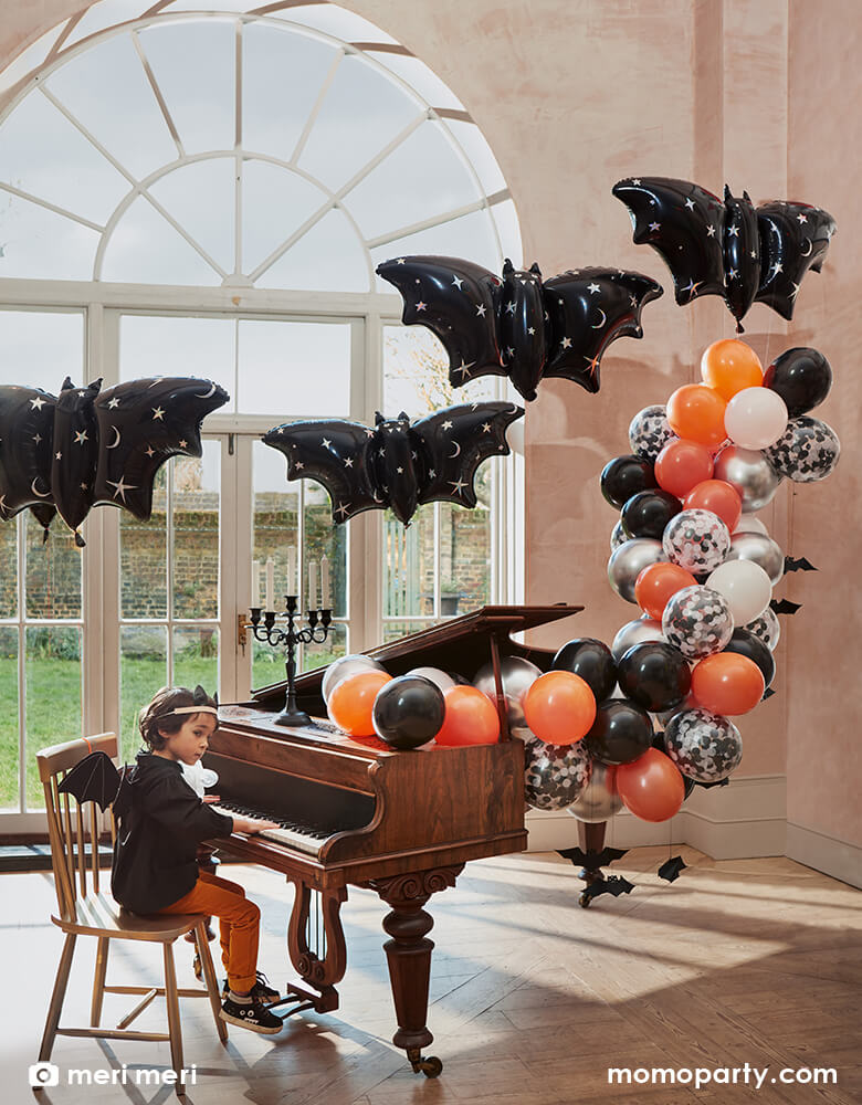A boy dressed in his bat costume sitting in front of a grand piano decorated with Momo Party's Halloween balloon garland in black, orange, silver, and white with bat decoration hanging, in the air there are four bat shaped foil balloons with silver moon and stars pattern on them, all creates a spooky yet chic inspiration for kid's Halloween party ideas.